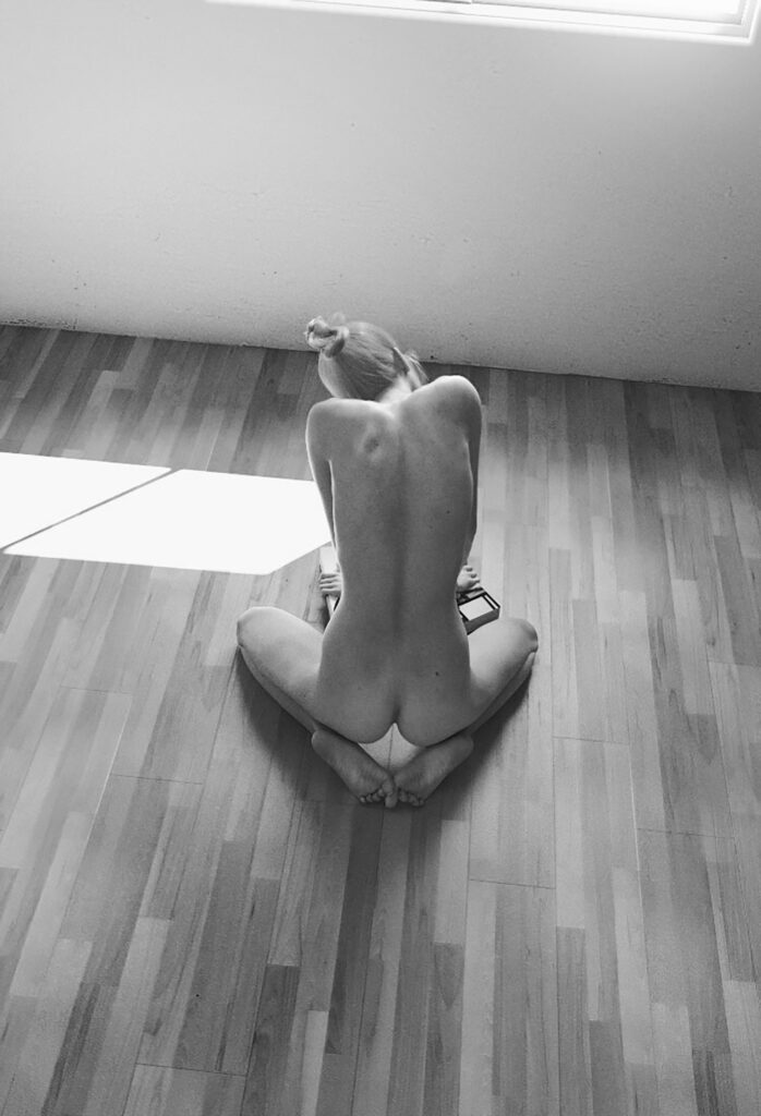 A woman with blonde hair tied in a bun sits nude on a wooden floor near a window with sunlight streaming in.